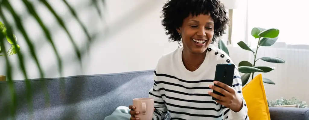 Eine Frau sitzt in einem Wohnzimmer auf dem Sofa und hält eine Tasse in der Hand. Sie blickt auf ihr Smartphone. Das Bild wird im Bereich "Besondere Versorgungsverträge | energie-Med|Portal" verwendet.