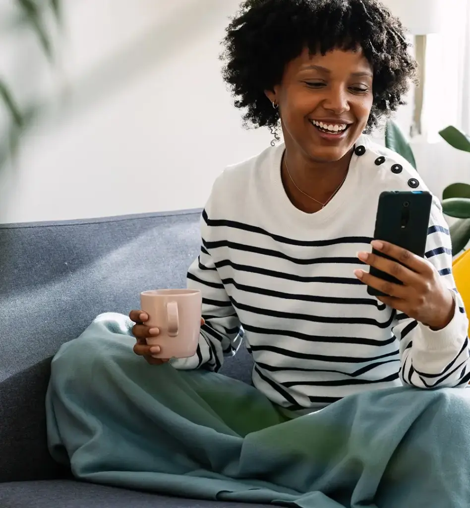 Eine Frau sitzt in einem Wohnzimmer auf dem Sofa und hält eine Tasse in der Hand. Sie blickt auf ihr Smartphone. Das Bild wird im Bereich "Besondere Versorgungsverträge | energie-Med|Portal" verwendet.