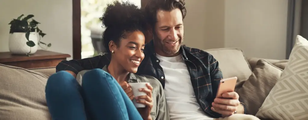 Ein Mann und eine Frau sitzt nebeneinander auf einer Couch. Er hält ein Smartphone in der Hand, auf das sie beide schauen. Sie hält eine Tasse in den Händen. Das Bild wird im Bereich "BudgetPlus Wahltarif – mehr für Sie drin!" verwendet.