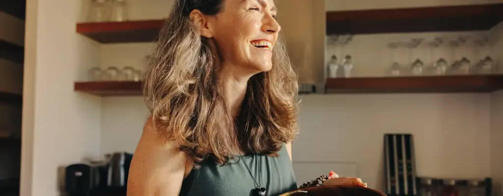 Eine Frau steht in der Küche, blickt aus dem Fenster. Die Sonne scheint in ihr Gesicht. Sie hält eine Holzschüssel mit Salat in den Händen. Das Bild wird im Bereich "califit - unser Ernährungs- und Bewegungscoaching" verwendet.