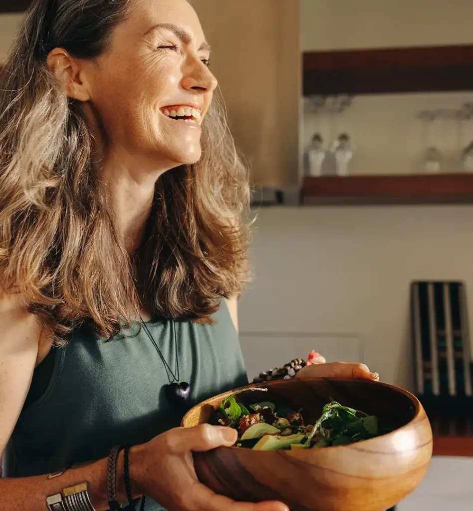 Eine Frau steht in der Küche, blickt aus dem Fenster. Die Sonne scheint in ihr Gesicht. Sie hält eine Holzschüssel mit Salat in den Händen. Das Bild wird im Bereich "califit - unser Ernährungs- und Bewegungscoaching" verwendet.