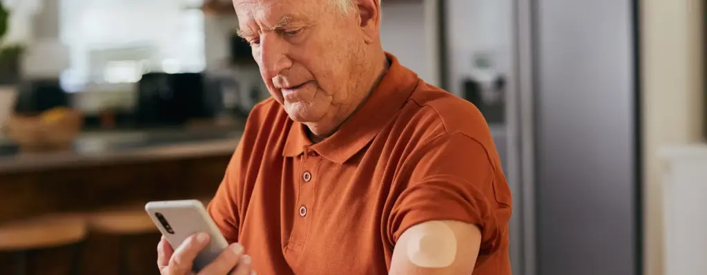 Ein älterer Mann schaut auf sein Smartphone. An seinem Oberarm sieht man einen Glukosemesser bei Diabetes. Das Bild wird im Bereich "Digitale Gesundheitsanwendungen – App auf Rezept (DiGAs)" verwendet.