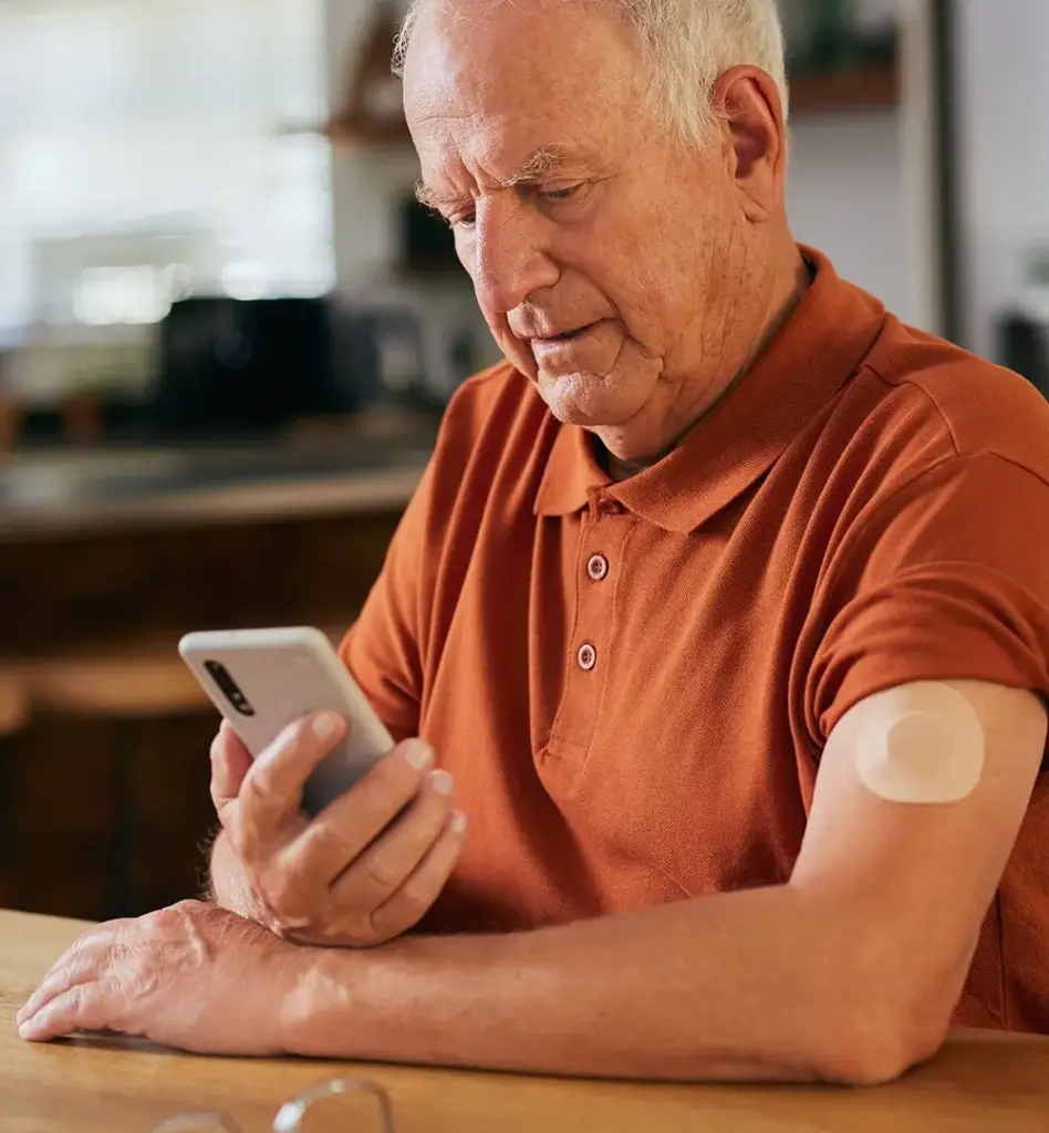 Ein älterer Mann schaut auf sein Smartphone. An seinem Oberarm sieht man einen Glukosemesser bei Diabetes. Das Bild wird im Bereich "Digitale Gesundheitsanwendungen – App auf Rezept (DiGAs)" verwendet.