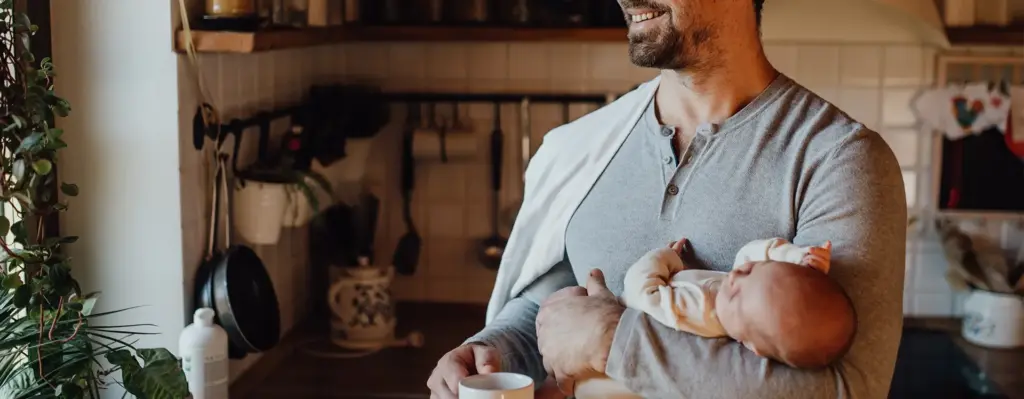 Ein Mann steht mit einem Baby im Arm in der Küche und blickt aus dem Fenster. Das Bild wird im Bereich "Elterngeld" verwendet.