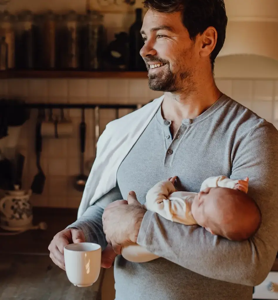 Ein Mann steht mit einem Baby im Arm in der Küche und blickt aus dem Fenster. Das Bild wird im Bereich "Elterngeld" verwendet.