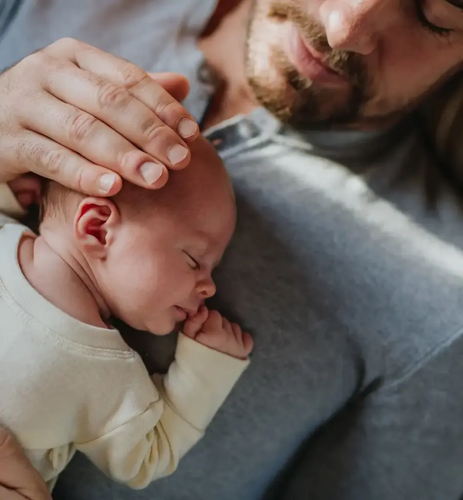 Ein Baby liegt auf dem Brustkorb des Vaters. Dieser hat seine Hand schützend auf den Kopf des Babys gelegt. Das Bild wird im Bereich "Elternzeit" verwendet.