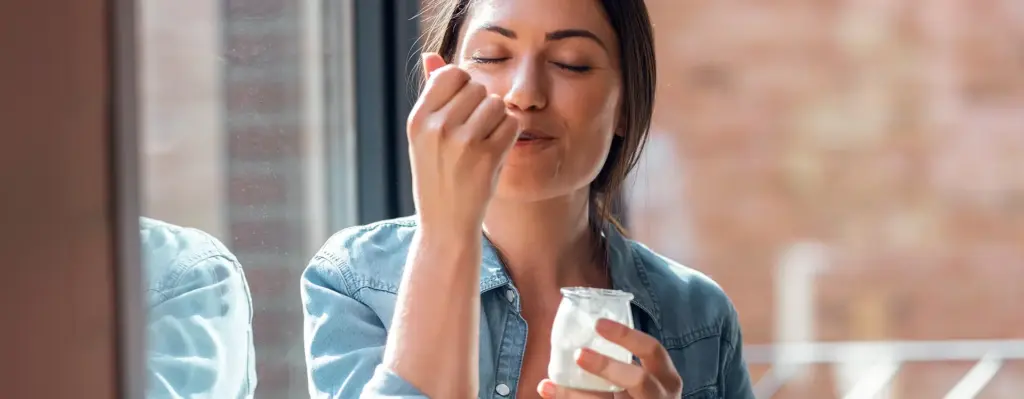Eine Frau sitzt an einem Fenster und isst aus einem Glas einen Joghurt. Sie hat genüsslich die Augen geschlossen. Das Bild wird im Bereich "Ernährungscoaching" verwendet.