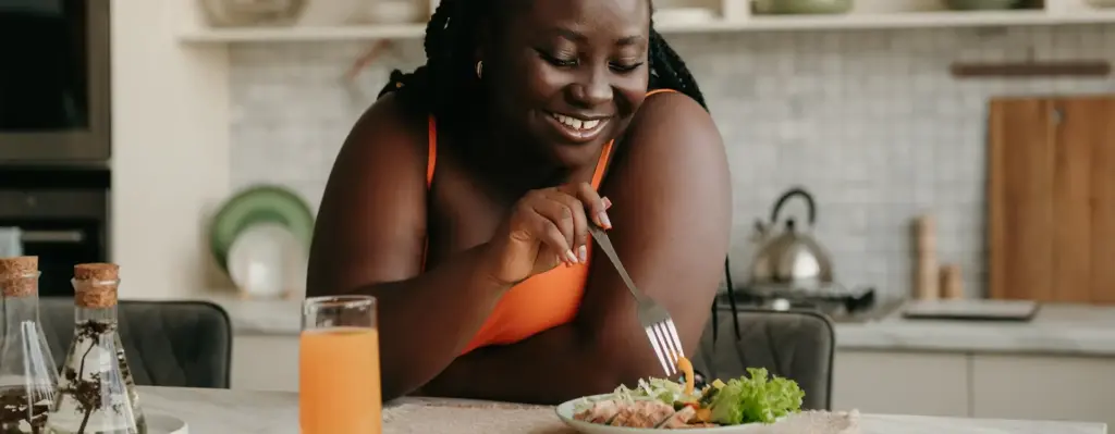 Eine Frau steht lachend in der Küche und piekt mit ihrer Gabel in einem Salat. Das Bild wird im Bereich "Ernährungsprogramm - RICHTIG ESSEN" verwendet.