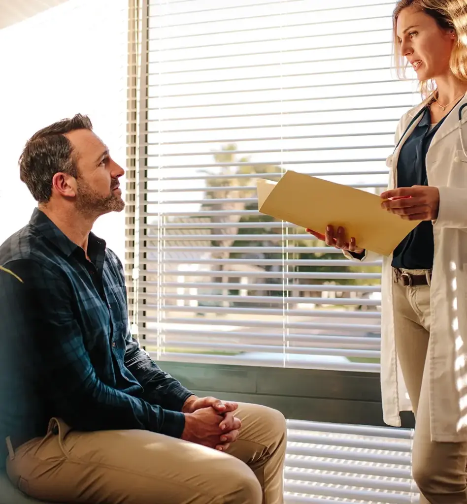 Eine Ärztin steht einem Patienten gegenüber, der auf einem Stuhl sitzt. Sie hält eine Mappe in der Hand. Das Bild wird im Bereich "Facharzt-Terminservice" verwendet.