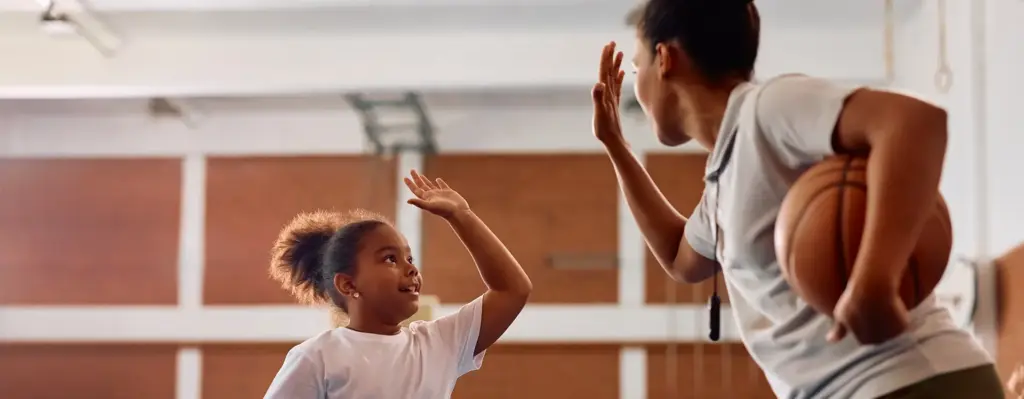 Eine Sportlehrerin gibt einem Mädchen in einer Turnhalle ein High Five. Sie hat einen Basketball in der Hand. Das Bild wird im Bereich "Fit von klein auf" verwendet.