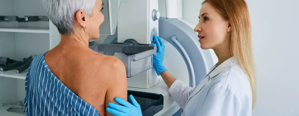 Eine ältere Frau steht an einem Röntgengerät für ein Mammographie Screening. Neben ihr steht eine Ärztin.