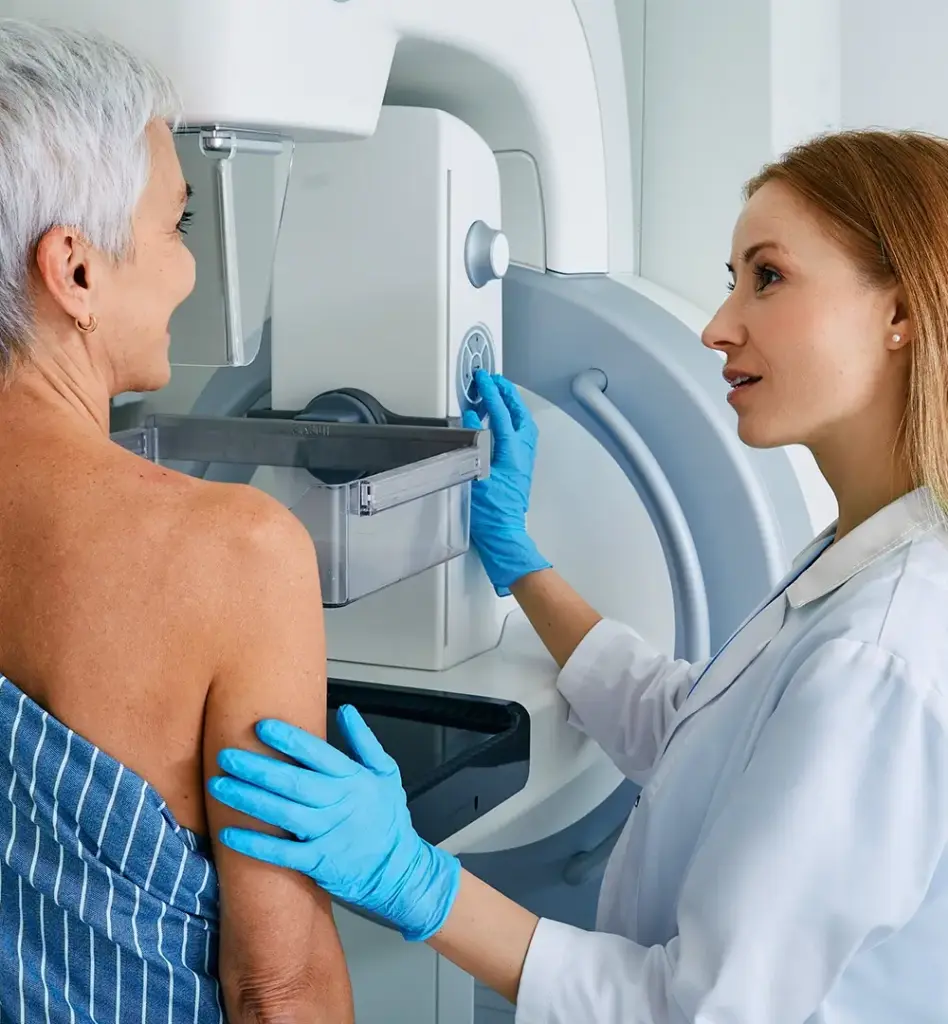 Eine ältere Frau steht an einem Röntgengerät für ein Mammographie Screening. Neben ihr steht eine Ärztin.