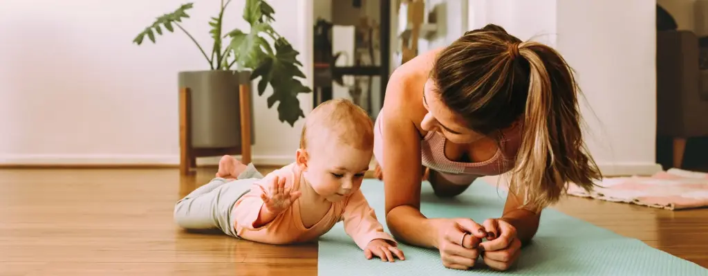 Eine Frau plankt auf einer Fitnessmatte. Neben ihr liegt ihr Baby in Bauchlage auf dem Boden. Das Bild wird im Bereich "Geburtsvorbereitung Rückbildungsgymnastik" verwendet.