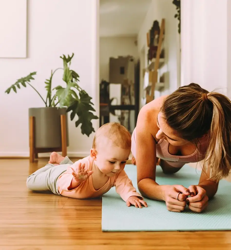 Eine Frau plankt auf einer Fitnessmatte. Neben ihr liegt ihr Baby in Bauchlage auf dem Boden. Das Bild wird im Bereich "Geburtsvorbereitung Rückbildungsgymnastik" verwendet.