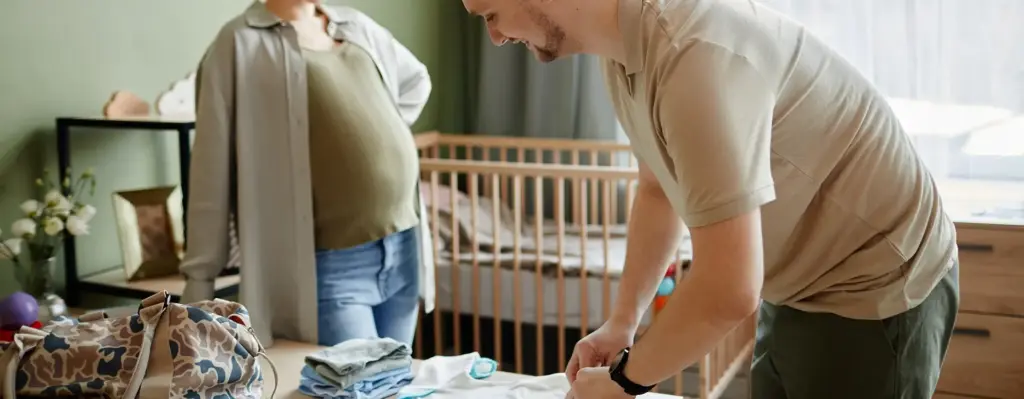 Das Bild zeigt einen Mann, der im Kinderzimmer Babykleidung faltet. Im Hintergrund steht eine schwangere Frau. Das Bild wird im Bereich "Geburtsvorbereitungskurse für beide Elternteile" verwendet.