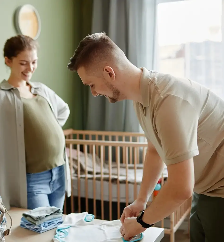 Das Bild zeigt einen Mann, der im Kinderzimmer Babykleidung faltet. Im Hintergrund steht eine schwangere Frau. Das Bild wird im Bereich "Geburtsvorbereitungskurse für beide Elternteile" verwendet.