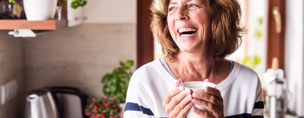 Eine lachende Frau steht mit einer Tasse in der Küche und blickt zur Seite. Das Bild wird im Bereich "Gesundheitskonto" verwendet.