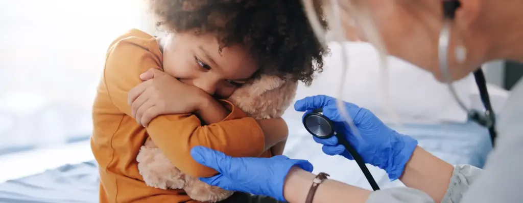 Das Bild zeigt ein Kleinkind, das verängstigt wirkt und sein Stofftier knuddelt. Eine Ärztin hält ein Stethoskop in der Hand und greift vorsichtig nach dem Arm. Das Bild wird im Bereich "Gesundheitsuntersuchungen für Kids" verwendet.