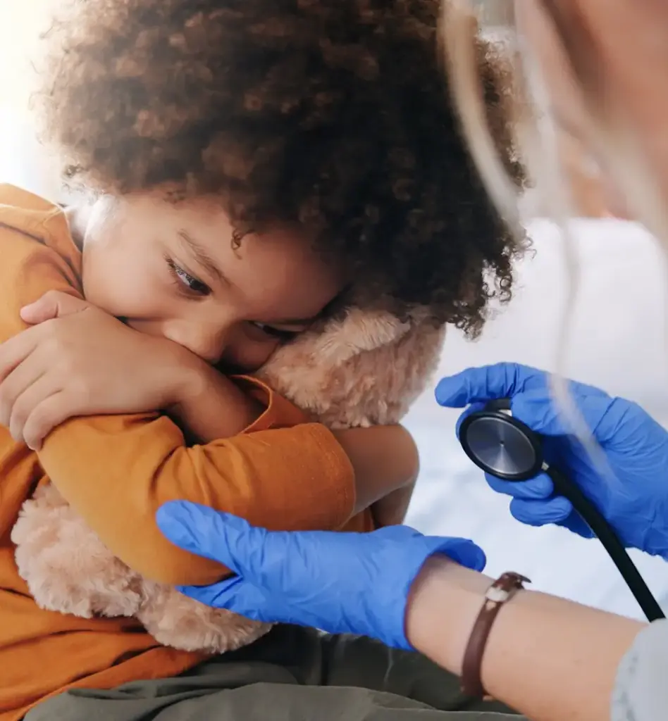 Das Bild zeigt ein Kleinkind, das verängstigt wirkt und sein Stofftier knuddelt. Eine Ärztin hält ein Stethoskop in der Hand und greift vorsichtig nach dem Arm. Das Bild wird im Bereich "Gesundheitsuntersuchungen für Kids" verwendet.