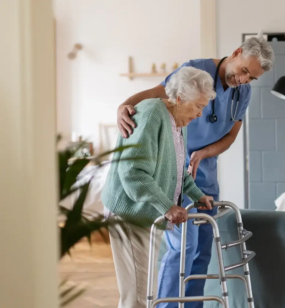 Das Bild zeigt eine ältere Frau mit Rollator beziehungsweiser einer Gehhilfe. Ein Pfleger läuft neben ihr. Das Bild wird im Bereich "Häusliche Krankenpflege" verwendet.