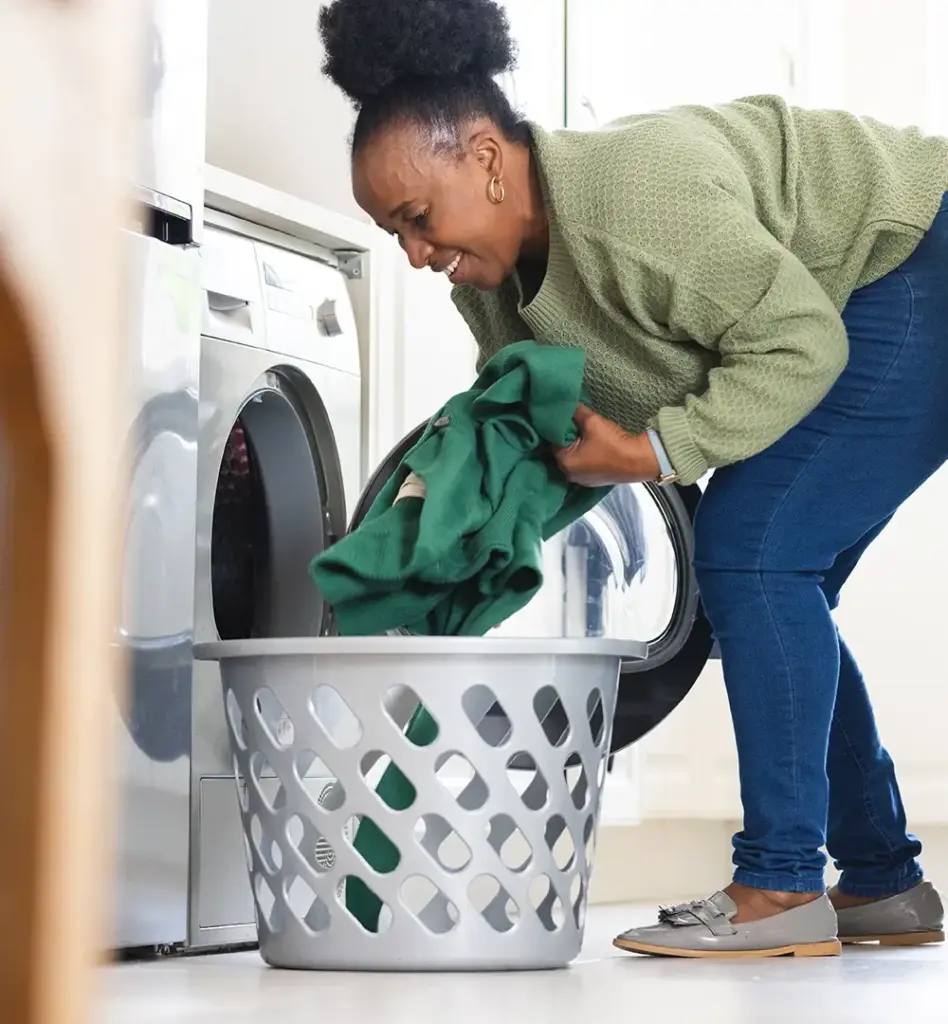 Eine Frau holt aus einem Wäschekorb Wäsche. Sie steht gebückt vor einer Waschmaschine. Das Bild wird im Bereich "Haushaltshilfe" verwendet.