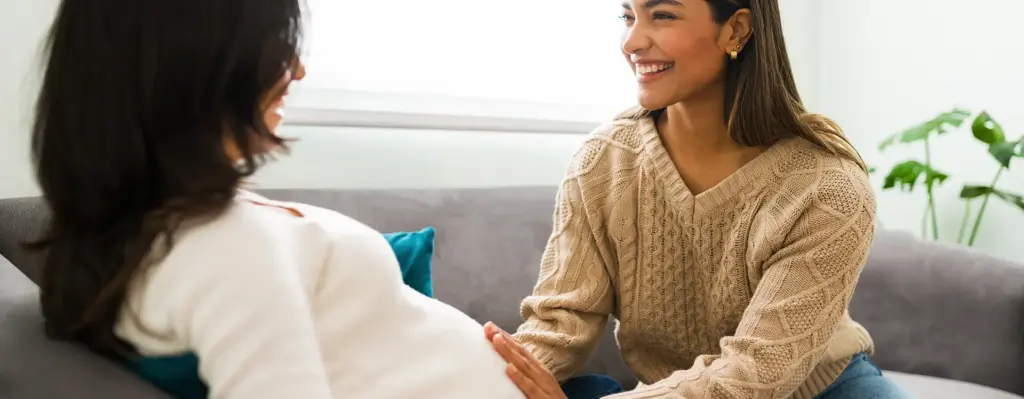 Eine schwangere Frau lehnt auf einem Sofa. Eine Frau tastet ihren Bauch ab. Das Bild wird im Bereich "Hebammenrufbereitschaft" verwendet.