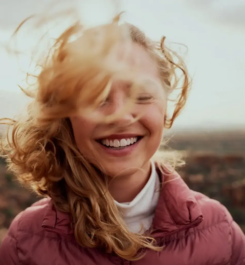 Eine lachende, blonde Frau mit geschlossenen Augen steht mit rosafarbener Jacke im Wind und ihre Haare wehen. Das Bild wird im Bereich "Homöopathie – Alternative Heilmethode" verwendet.