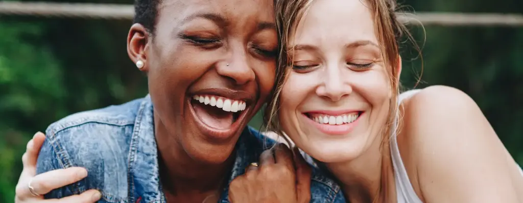 Zwei Frauen lachen mit geschlossenen Augen und umarmen sich nebeneinandersitzend. Das Bild wird im Bereich "Implantate" verwendet.
