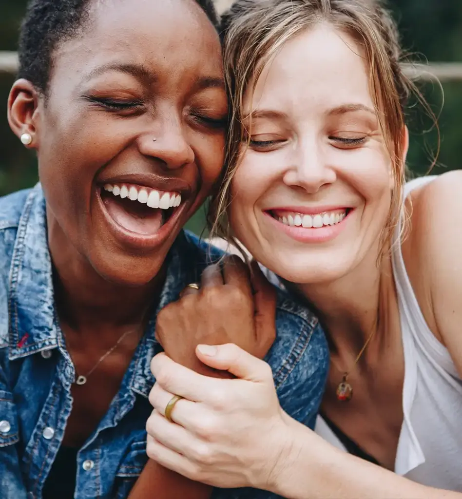 Zwei Frauen lachen mit geschlossenen Augen und umarmen sich nebeneinandersitzend. Das Bild wird im Bereich "Implantate" verwendet.