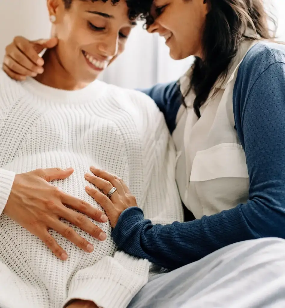 Zwei Frauen sind einander zugewandt. Die eine Frau fasst der anderen an den Bauch. Die andere ist schwanger. Beide lächeln. Das Bild wird im Bereich "Keleya Schwangerschaftsapp" verwendet.
