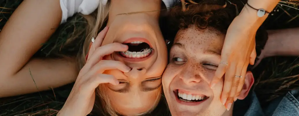 Ein Teenager und eine Teenagerin liegen Kopf an Kopf auf einer Wiese und lachen. Das Mädchen trägt eine Zahnspange. Das Bild wird im Bereich "Kieferorthopädische Behandlung" verwendet.