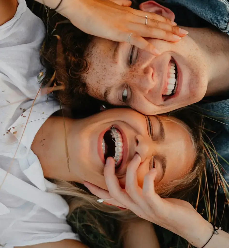 Ein Teenager und eine Teenagerin liegen Kopf an Kopf auf einer Wiese und lachen. Das Mädchen trägt eine Zahnspange. Das Bild wird im Bereich "Kieferorthopädische Behandlung" verwendet.