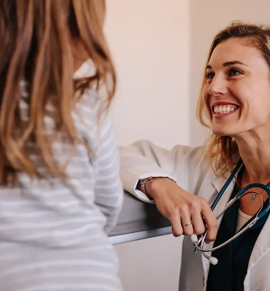 Eine Ärztin kniet neben einem Mädchen und lächelt es an. Um den Hals hat sie ein Stethoskop.