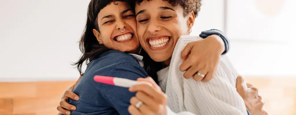 Zwei Frauen umarmen sich und strahlen vor Glück. Eine von ihnen hält einen Schwangerschaftstest in der Hand. Das Bild wird im Bereich "BKK Kinderwunsch" verwendet.