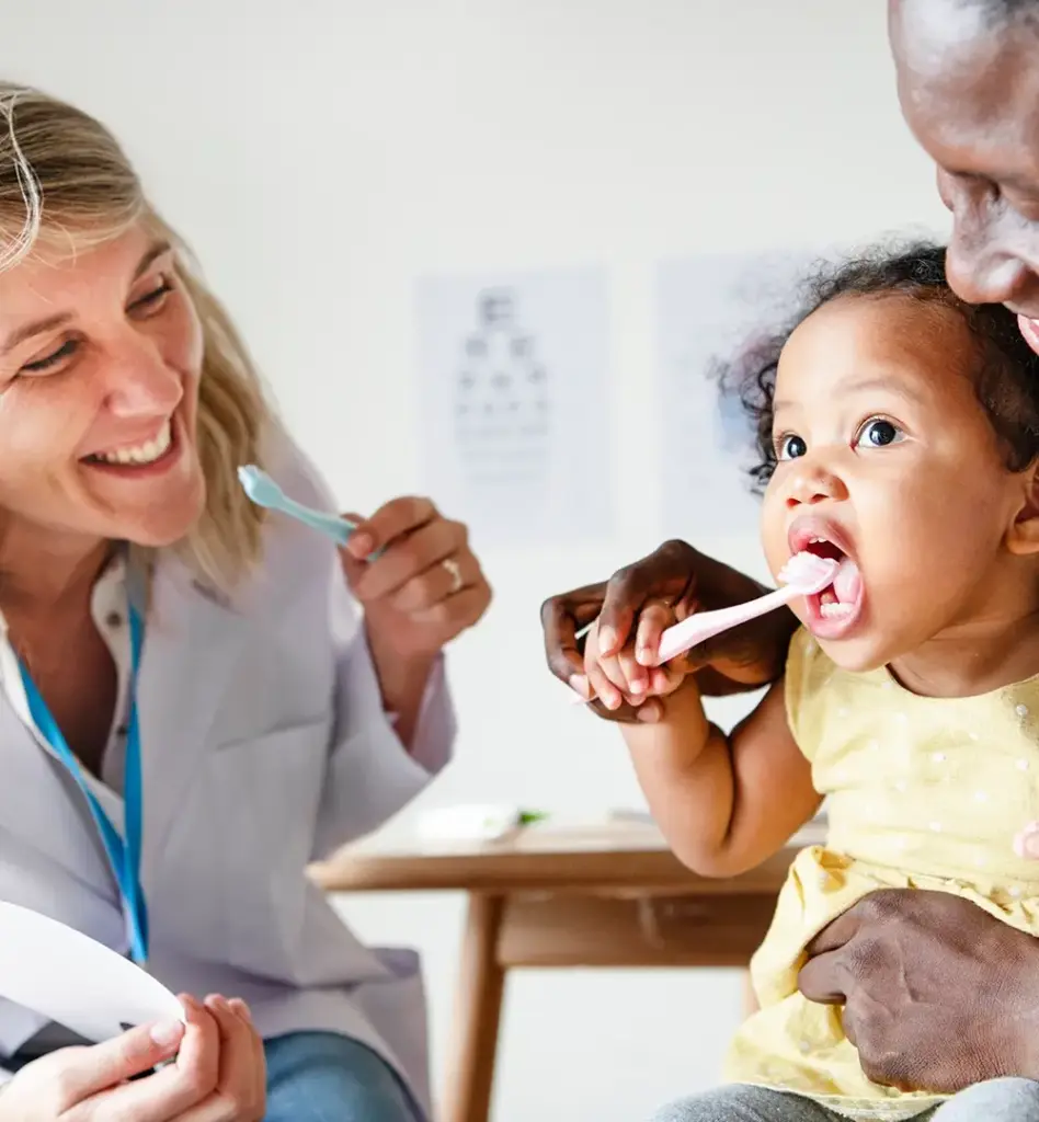 Das Bild zeigt eine Ärztin, die eine Zahnbürste in der Hand hat und einem Mädchen, das auf dem Schoß ihrer Mama sitzt vormacht, wie man die Zähne putzt. Das Mädchen hat ebenfalls eine Zahnbürste in der Hand. Das Bild wird im Bereich "Kinderzahnprophylaxe" verwendet.