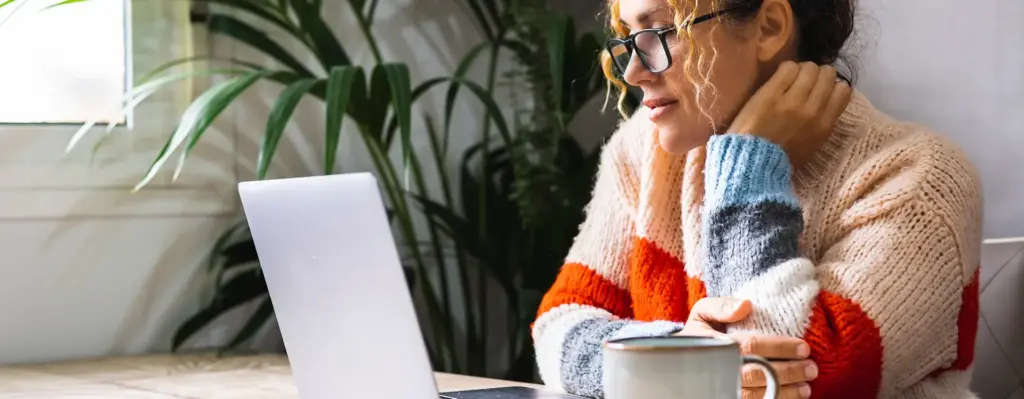 Eine Frau sitzt an einem Tisch vor einem Laptop. Sie trägt eine Brille. Auf dem Tisch steht noch eine Tasse. Das Bild wird im Bereich "Klinikfinder" verwendet.