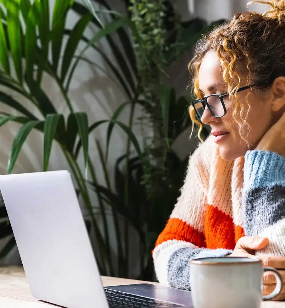 Eine Frau sitzt an einem Tisch vor einem Laptop. Sie trägt eine Brille. Auf dem Tisch steht noch eine Tasse. Das Bild wird im Bereich "Klinikfinder" verwendet.