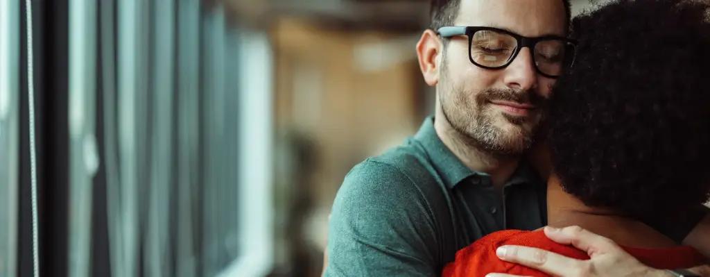 Ein junger Mann mit Brille umarmt eine Frau. Diese ist nur von hinten zu sehen. Das Bild wird im Bereich "Kraft zu Leben – Unterstützung für Krebskranke" verwendet.
