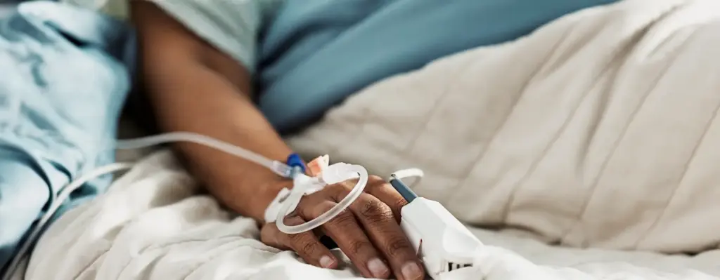 Man sieht eine Nahaufnahme eines Armes, einer Person im Krankenhausbett, die einen Katheterzugang gelegt hat und einen Vitalwertmessgerät am Finger hat. Das Bild wird im Bereich "Krankenhausbehandlung" verwendet.