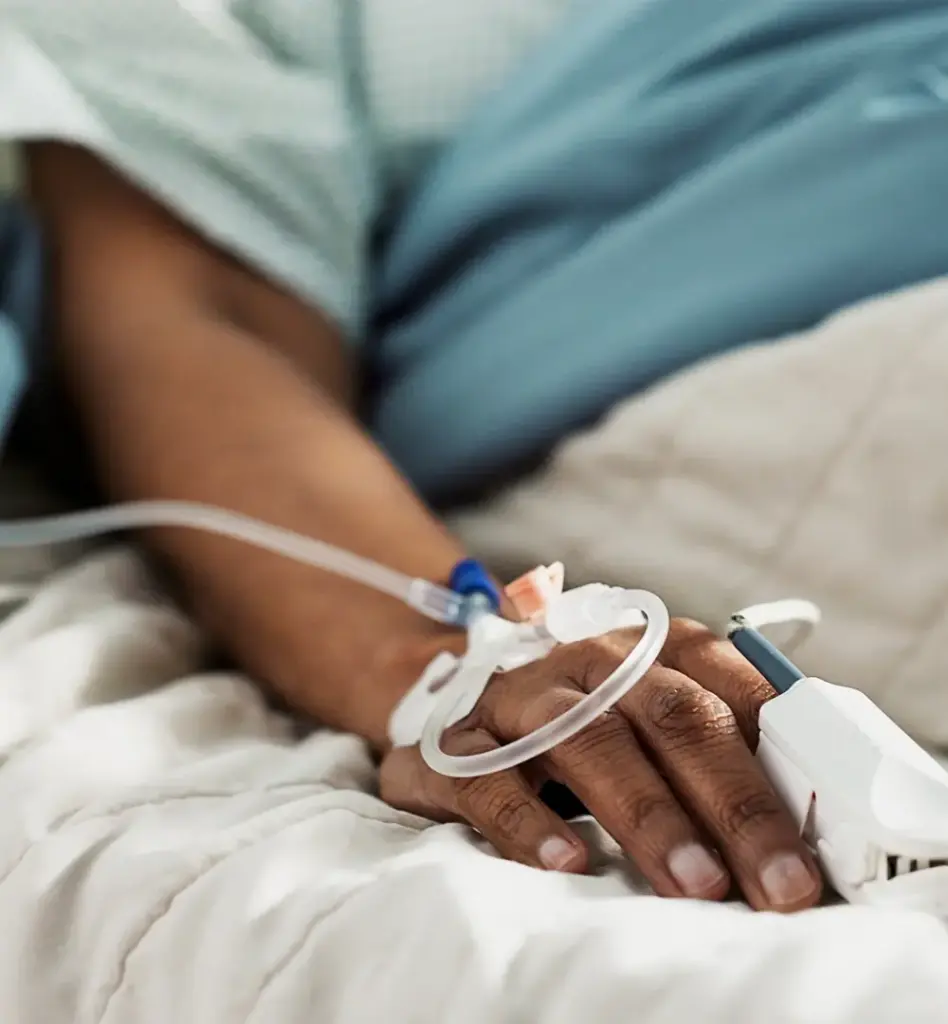 Man sieht eine Nahaufnahme eines Armes, einer Person im Krankenhausbett, die einen Katheterzugang gelegt hat und einen Vitalwertmessgerät am Finger hat. Das Bild wird im Bereich "Krankenhausbehandlung" verwendet.