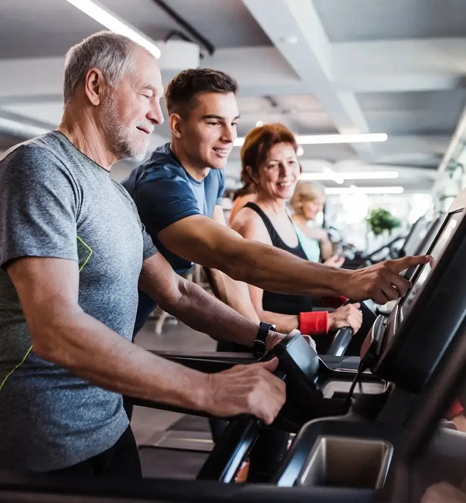 Ein älterer Mann steht auf einem Laufband im Fitnessstudio. Ein junger Mann steht daneben und tippt auf dem Bildschirm des Laufbandes. Im Hintergrund sieht man noch Frauen auf dem Laufband. Das Bild wird im Bereich "Kuren" verwendet.