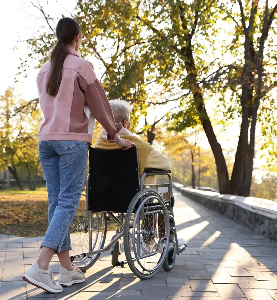 Eine ältere Frau sitzt im Rollstuhl. Eine jüngere Frau schiebt sie einen Weg entlang. Das Bild wird im Bereich "Kurzzeitpflege bei fehlender Pflegebedürftigkeit" verwendet.