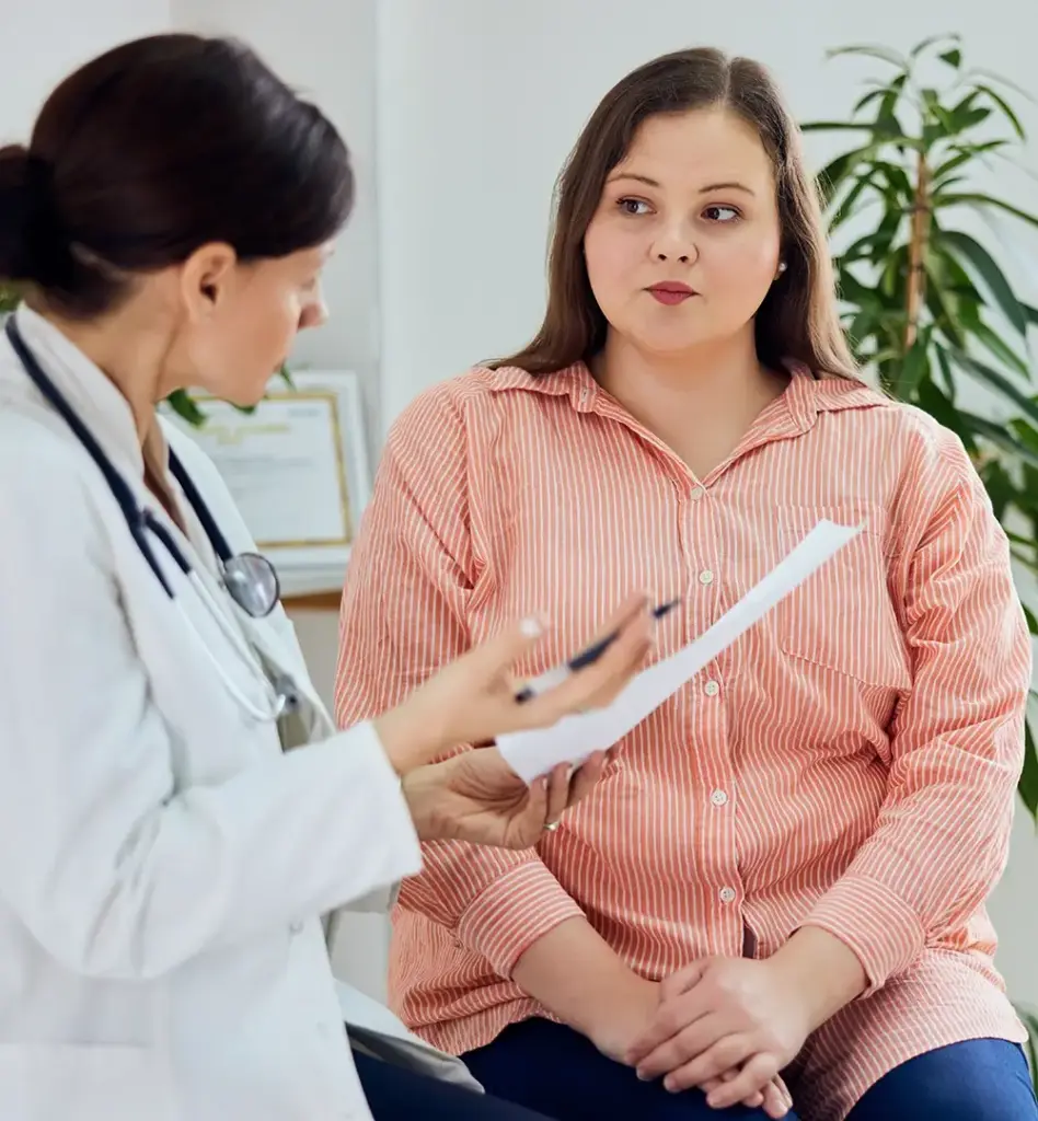 Das Bild zeigt eine Ärztin, die einen Zettel und Kugelschreiber in den Händen hält und gestikuliert. Sie erklärt einer Patientin, die auf der Behandlungsliege sitzt etwas. Das Bild wird im Bereich "Lasertherapie bei vaginalen-Erkrankungen" verwendet.