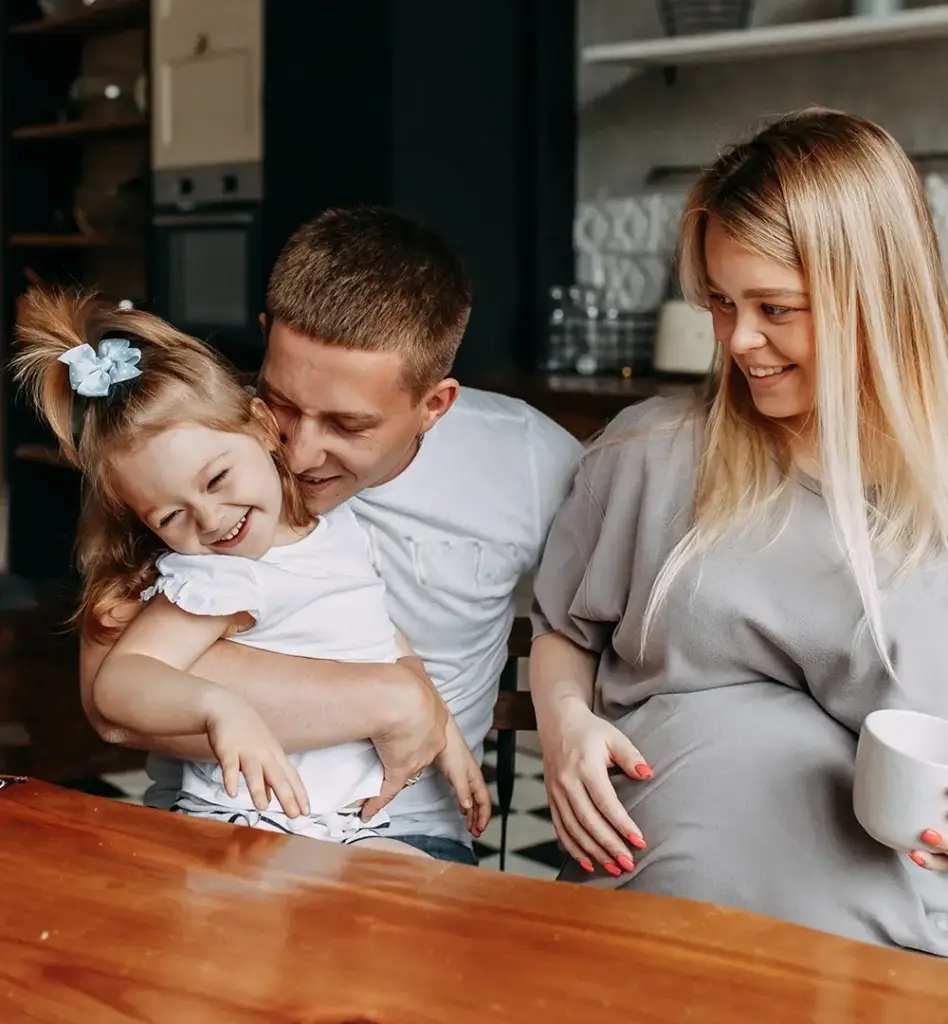 Ein Mädchen sitzt auf dem Schoß eines Mannes. Sie lachen und kuscheln. Neben ihnen sitzt eine schwangere Frau. Sie befinden sich in einer Küche an einem Esstisch. Das Bild wird im Bereich "Leistungen für Familie und Schwangerschaft" verwendet.