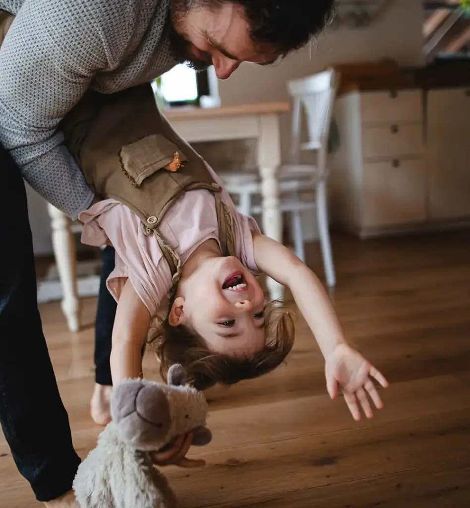 Ein Mann hält ein kleines Mädchen auf dem Arm und schwenkt sie lachend kopfüber nach unten. Das Mädchen lacht und hält ein Stofftier in der Hand. Das Bild wird im Bereich "Family Plus" verwendet.