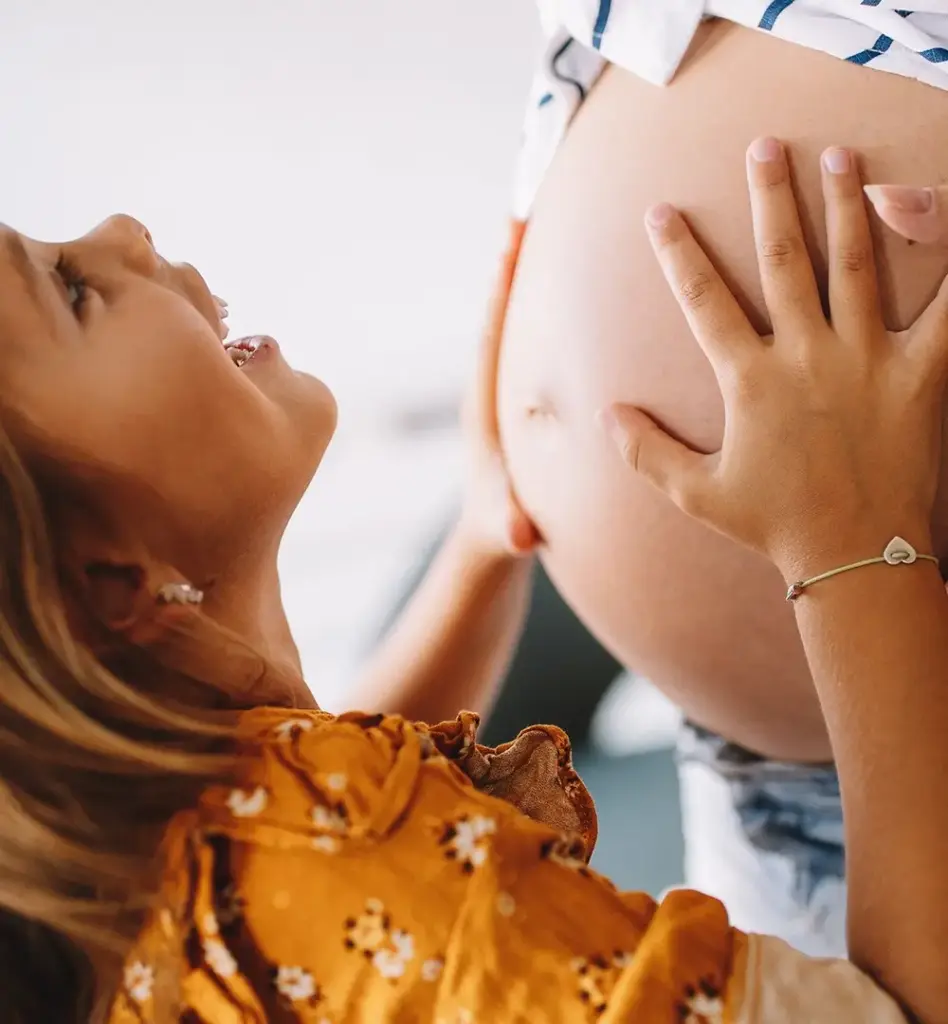 Das Bild zeigt ein kleines Mädchen, das strahlend den Bauch ihrer schwangeren Mama anfasst. Das Mädchen blickt nach oben zur Mutter. Von der Mutter sieht man nur den Bauch. Das Bild wird im Bereich "Mamabonus Babybonus" verwendet.