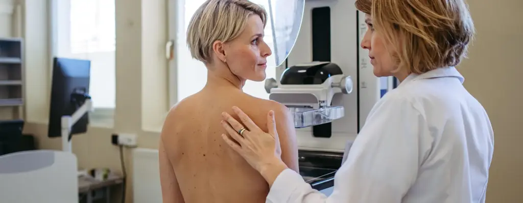 Eine Frau mit kurzen blonden Haaren steht an einem Röntgengerät für ein Mammographie-Screening. Eine Ärztin steht neben ihr und hat ihre Hand auf das Schulterblatt der Frau gelegt. Das Bild wird im Bereich "Mammographie-Screening" verwendet.