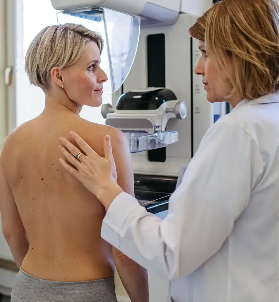 Eine Frau mit kurzen blonden Haaren steht an einem Röntgengerät für ein Mammographie-Screening. Eine Ärztin steht neben ihr und hat ihre Hand auf das Schulterblatt der Frau gelegt. Das Bild wird im Bereich "Mammographie-Screening" verwendet.