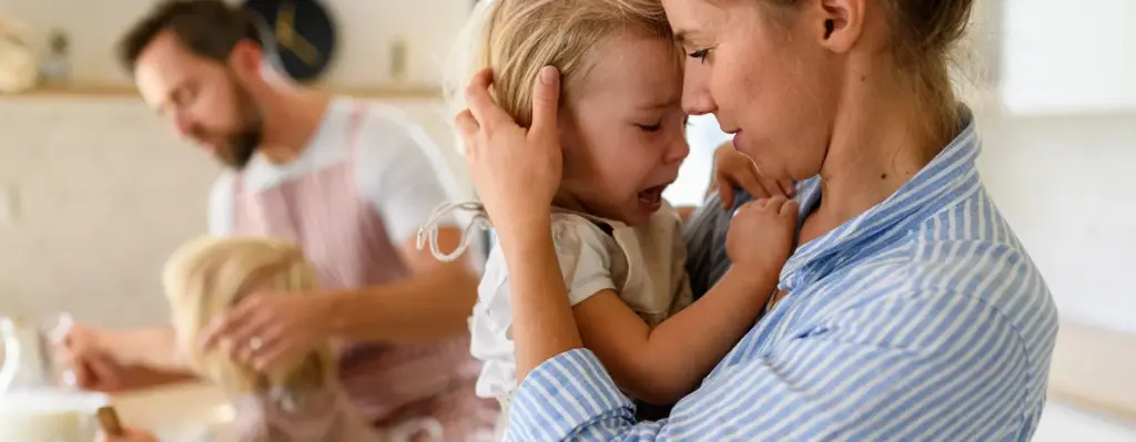 Das Bild zeigt eine Frau in Bluse, die ein kleines Mädchen auf dem Arm hat. Das Mädchen weint und die Frau tröstet es. Das Bild wird im Bereich "Mapadoo Onlinekurse für Familien" verwendet.