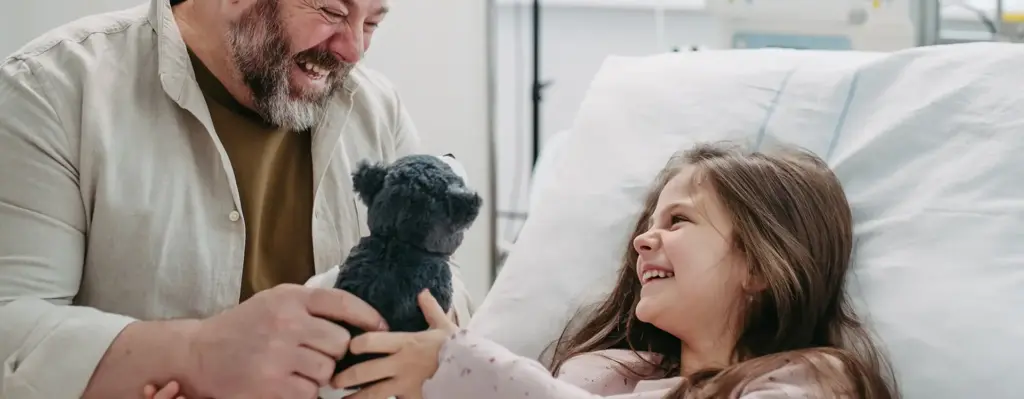Das Bild zeigt ein Mädchen, dass in einem Krankenhausbett liegt. Neben ihr sitzt ein Mann - ihr Vater. Er gibt seiner Tochter ein Kuscheltier. Beide lachen. Das Bild wird im Bereich "Mitaufnahme der Eltern beim Krankenhausaufenthalt eines Kindes" verwendet.